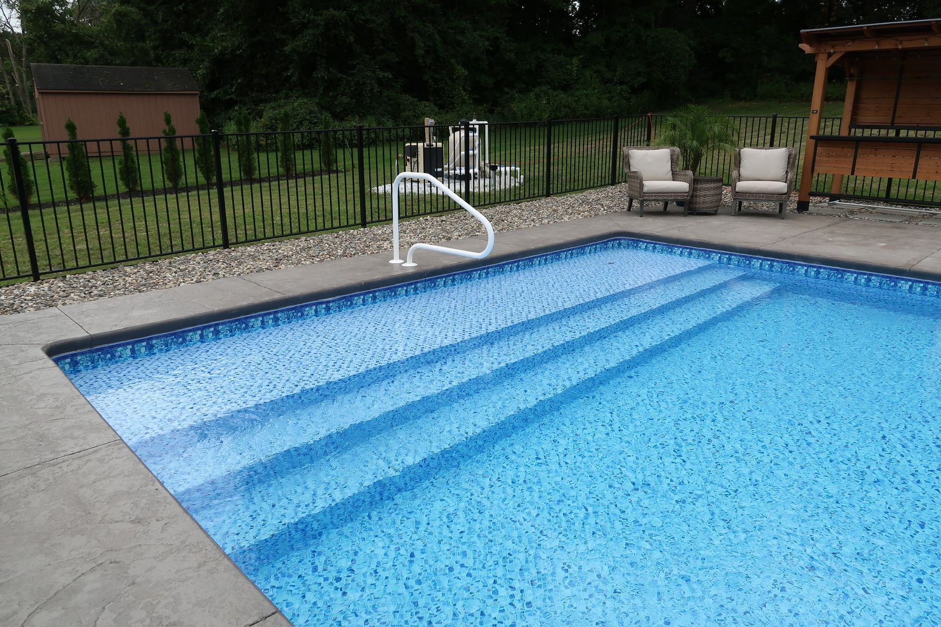 Pool with steps, handrail, and lounge chairs next to a fence and landscaping.