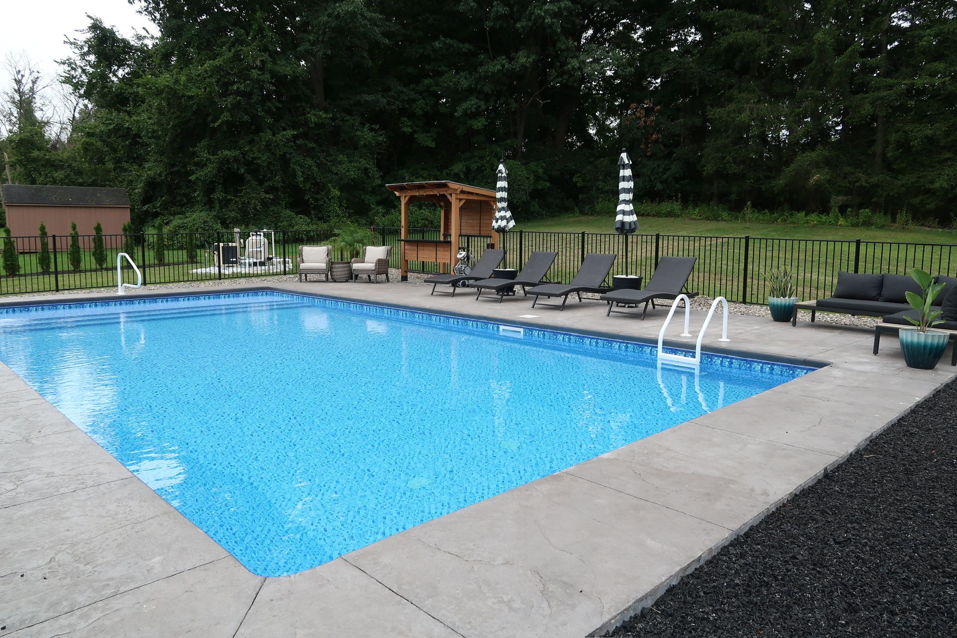 Rectangular pool with concrete surround; lounge chairs, pergola, and green trees.