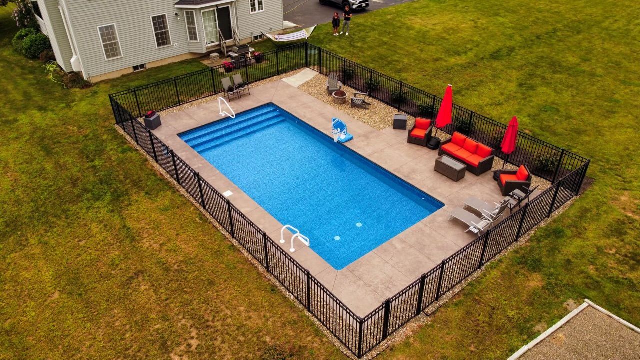 An aerial view of a rectangular blue swimming pool surrounded by a black fence, with patio furniture.