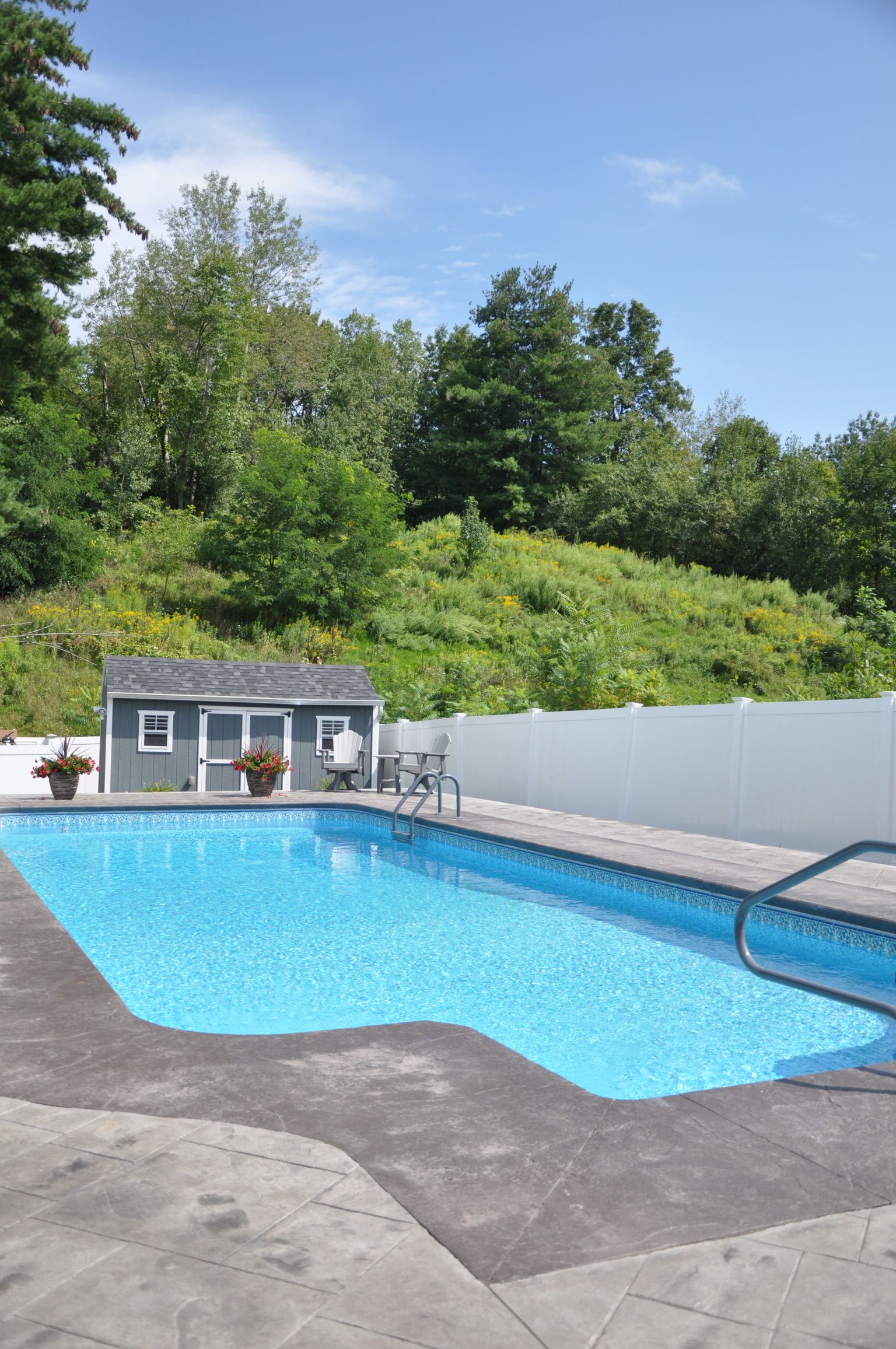 Swimming pool with blue water, gray patio, white fence, small shed, and trees on a hill.