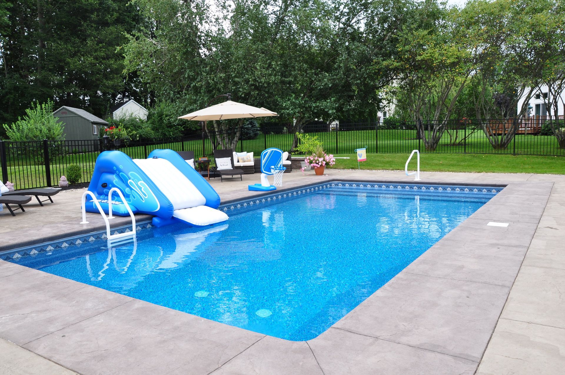 Rectangular blue swimming pool with inflatable slide, patio, and green backyard.