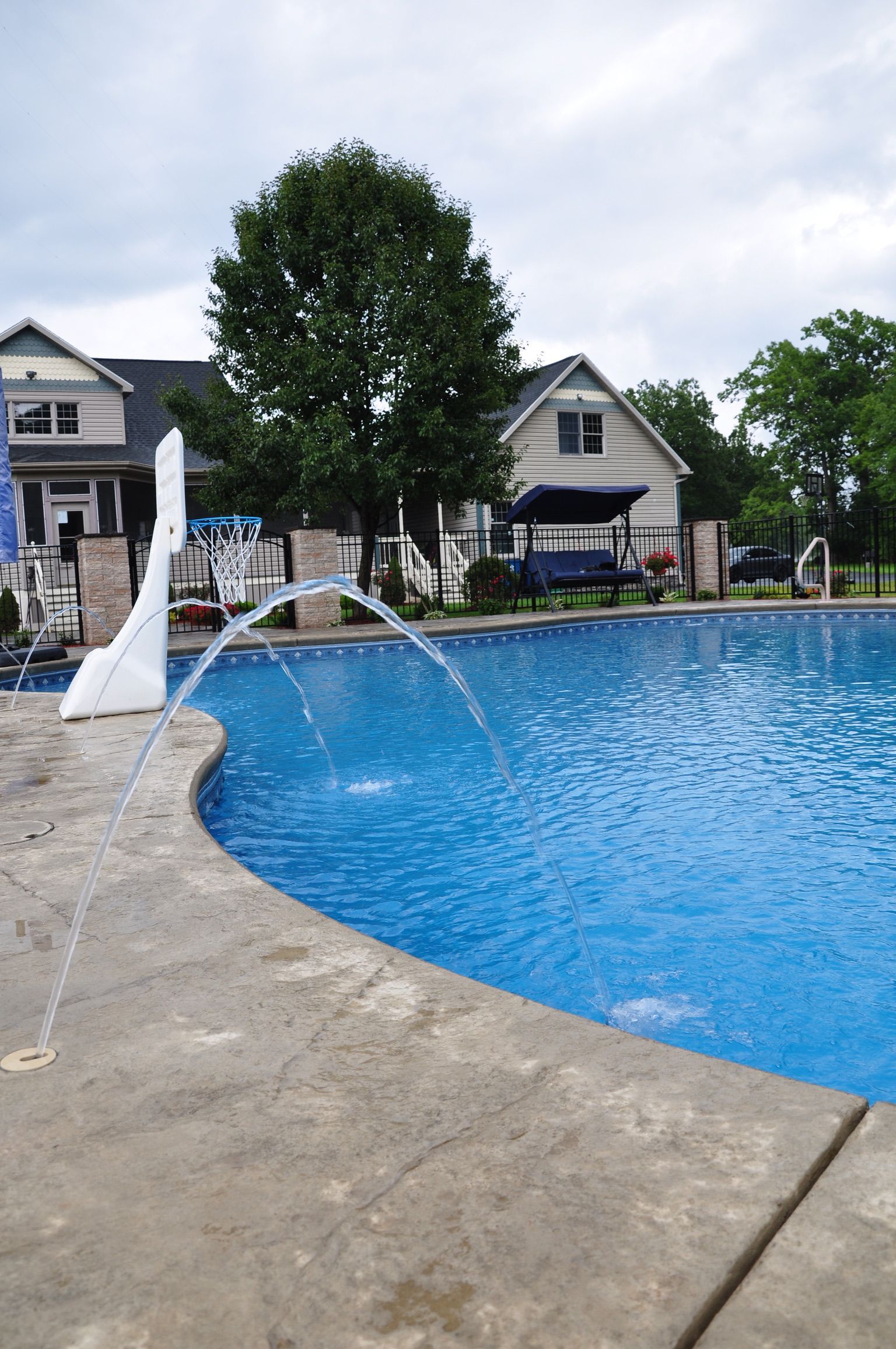Swimming pool with water fountain; houses and trees in background.
