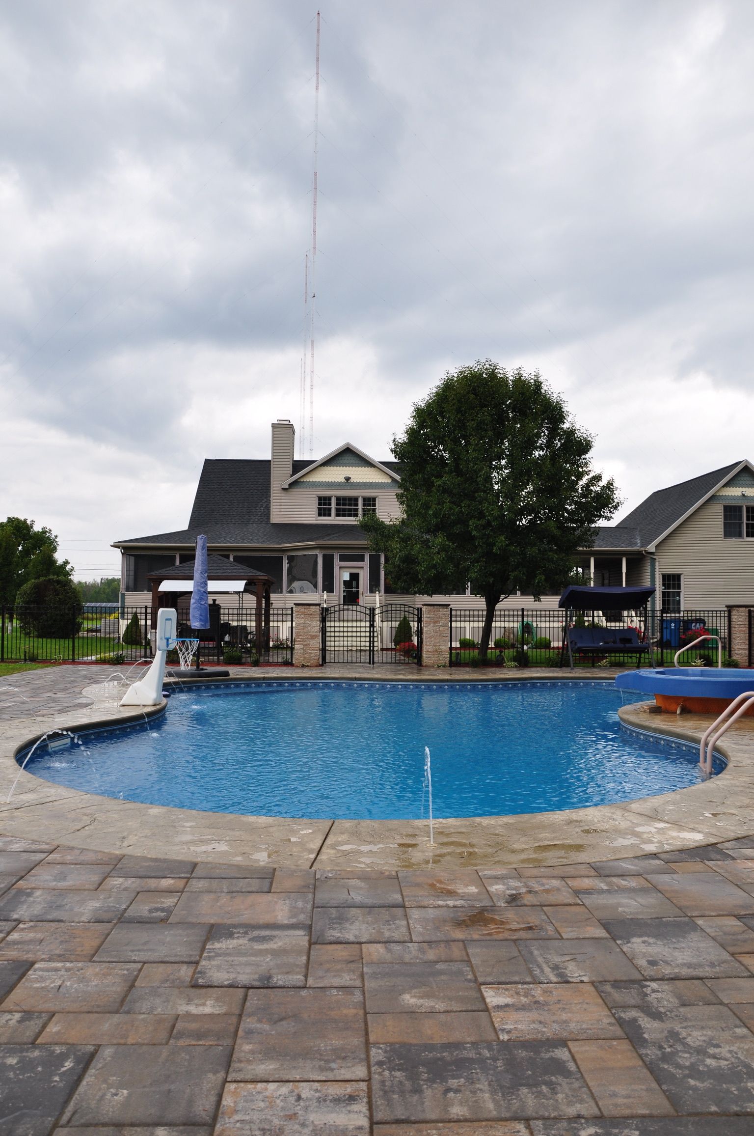 Backyard pool with stone patio, large house, tree, and overcast sky.