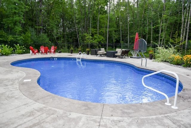 Swimming pool with blue water and concrete deck, surrounded by trees and patio furniture.