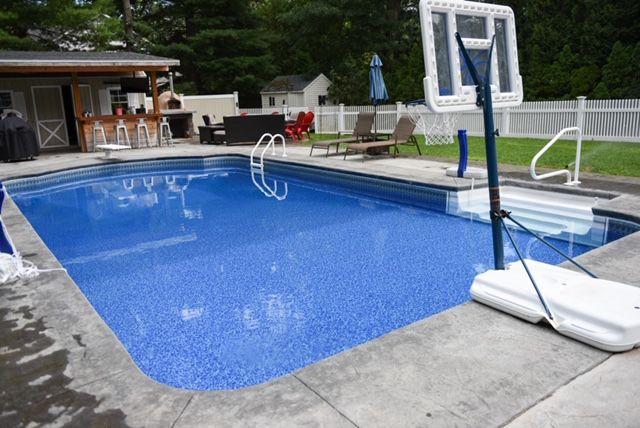 Rectangular pool with blue water and basketball hoop, concrete deck, backyard setting.