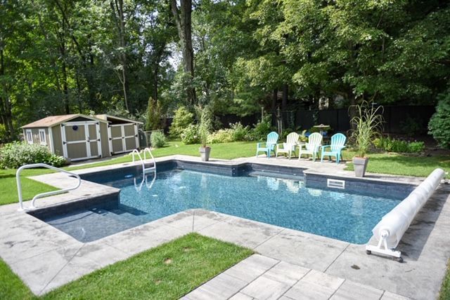 Backyard pool with stone patio, green chairs, and shed surrounded by trees.