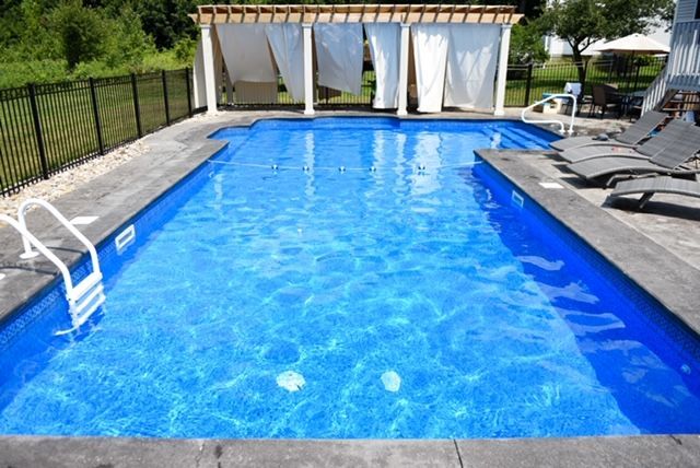 Rectangular blue swimming pool with white curtains, a black fence, and lounge chairs.