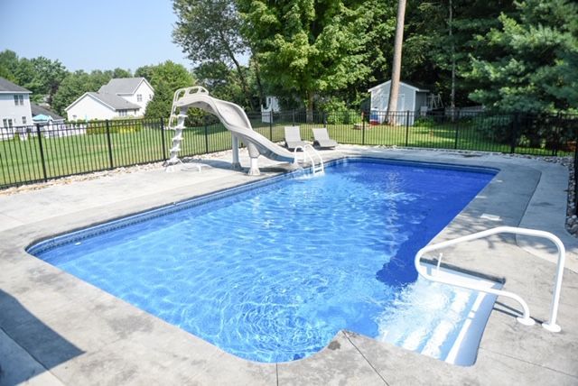 Rectangular in-ground pool with slide, surrounded by concrete, fence, and green trees.