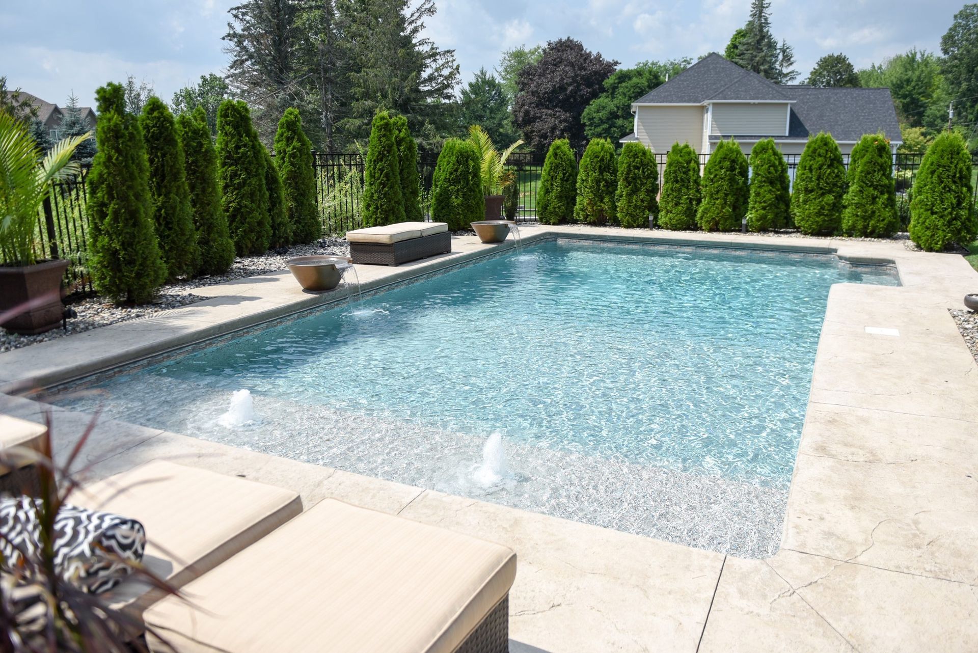 A rectangular swimming pool surrounded by stone patio, greenery, and a house. Two lounge chairs in foreground.