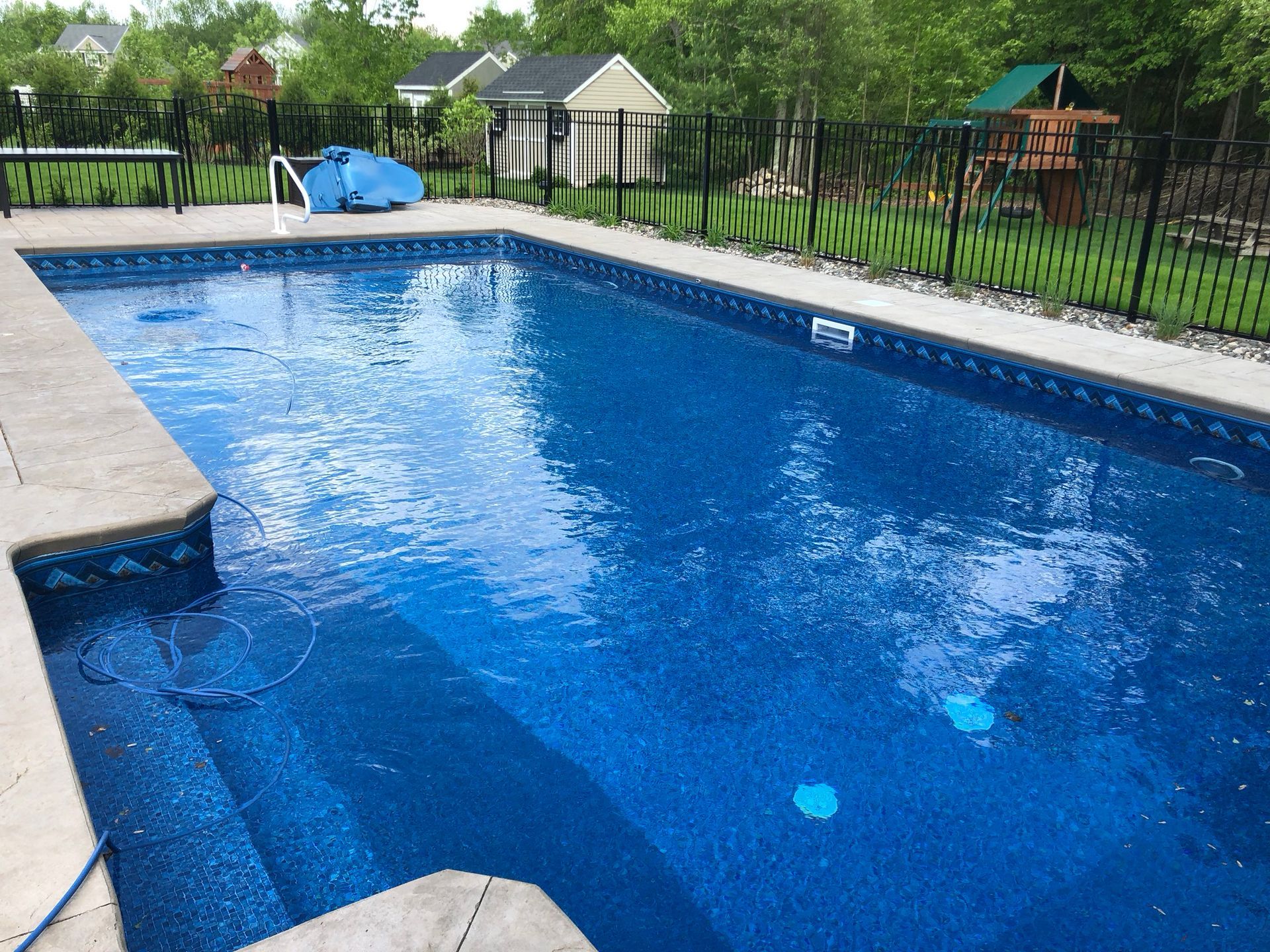 A rectangular blue swimming pool surrounded by concrete and a black fence, with buildings and trees in the background.