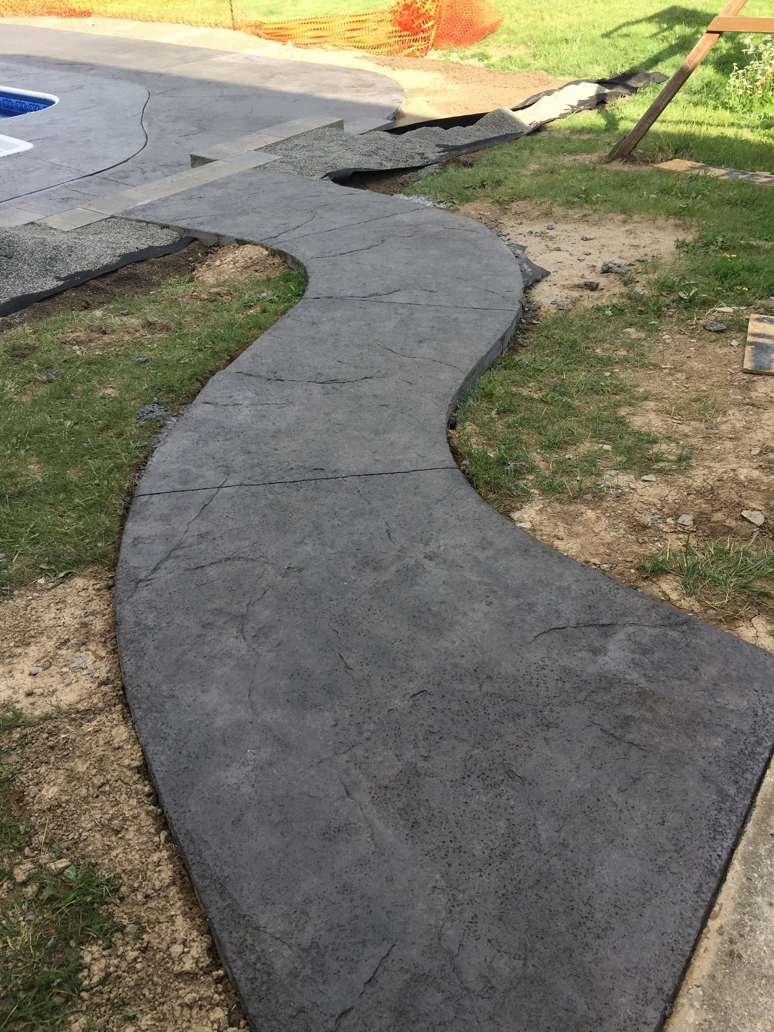 Curving concrete pathway through grass, leading towards a pool. Dark gray color.