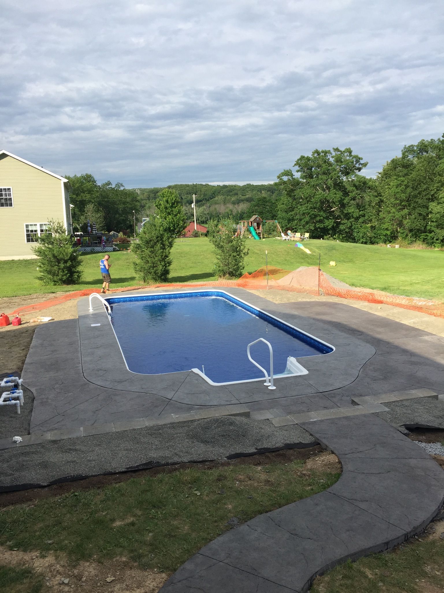 Rectangular swimming pool surrounded by dark gray concrete. Person standing near the pool on green lawn, under cloudy sky.