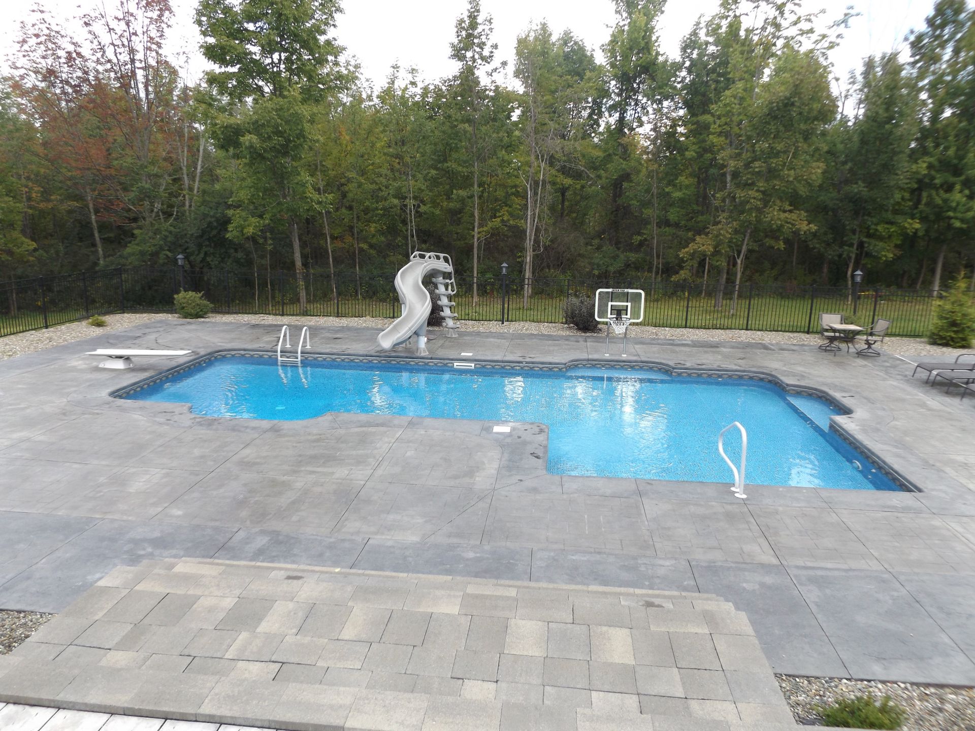 Backyard pool with a slide, basketball hoop, and trees. Blue water, gray patio.