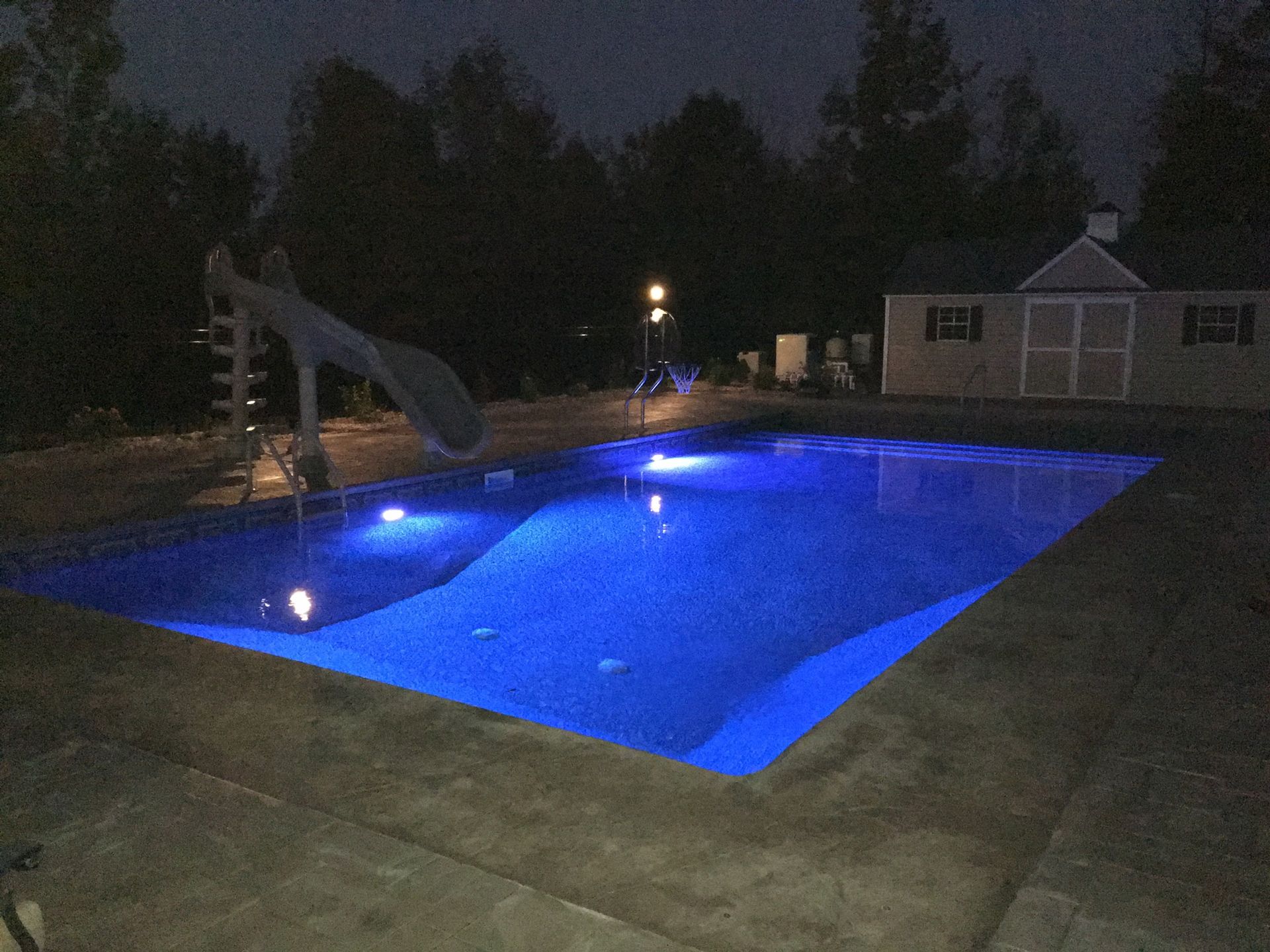 A nighttime view of a rectangular swimming pool lit with blue lights, with a slide and a shed in the background.