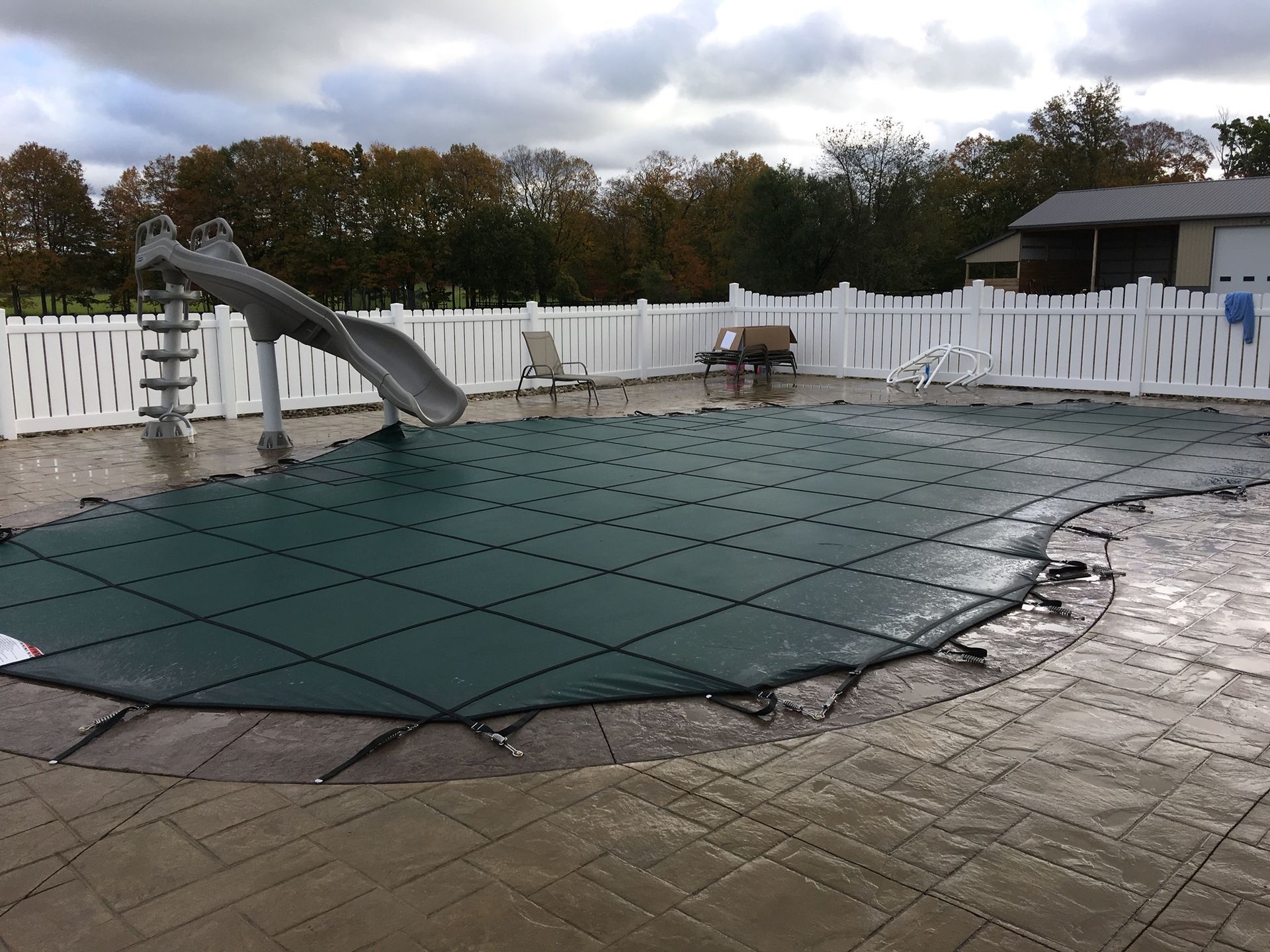 Swimming pool with green cover, slide, surrounded by a white fence on a cloudy day.