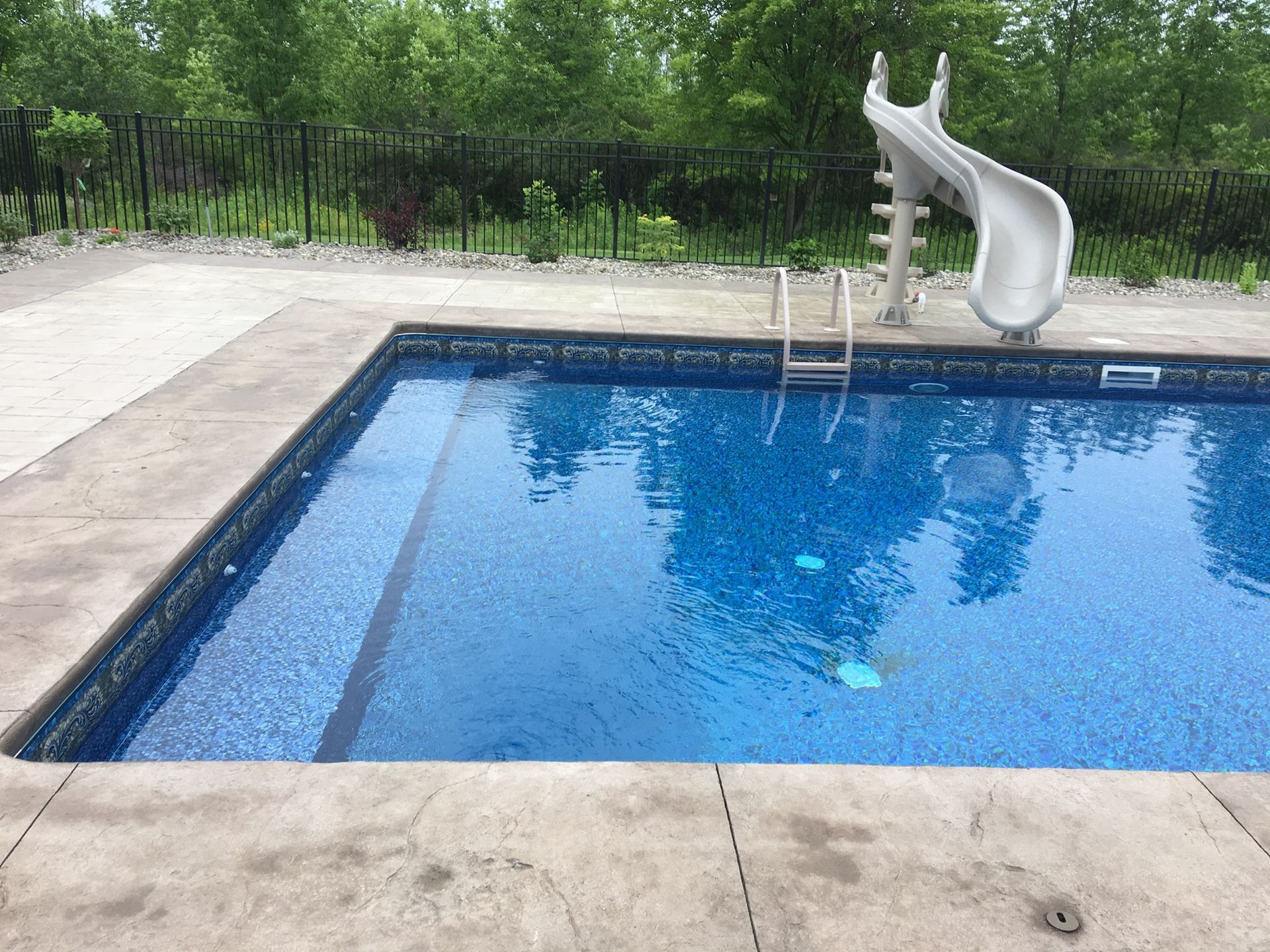 Swimming pool with a water slide and ladder, surrounded by concrete and greenery.