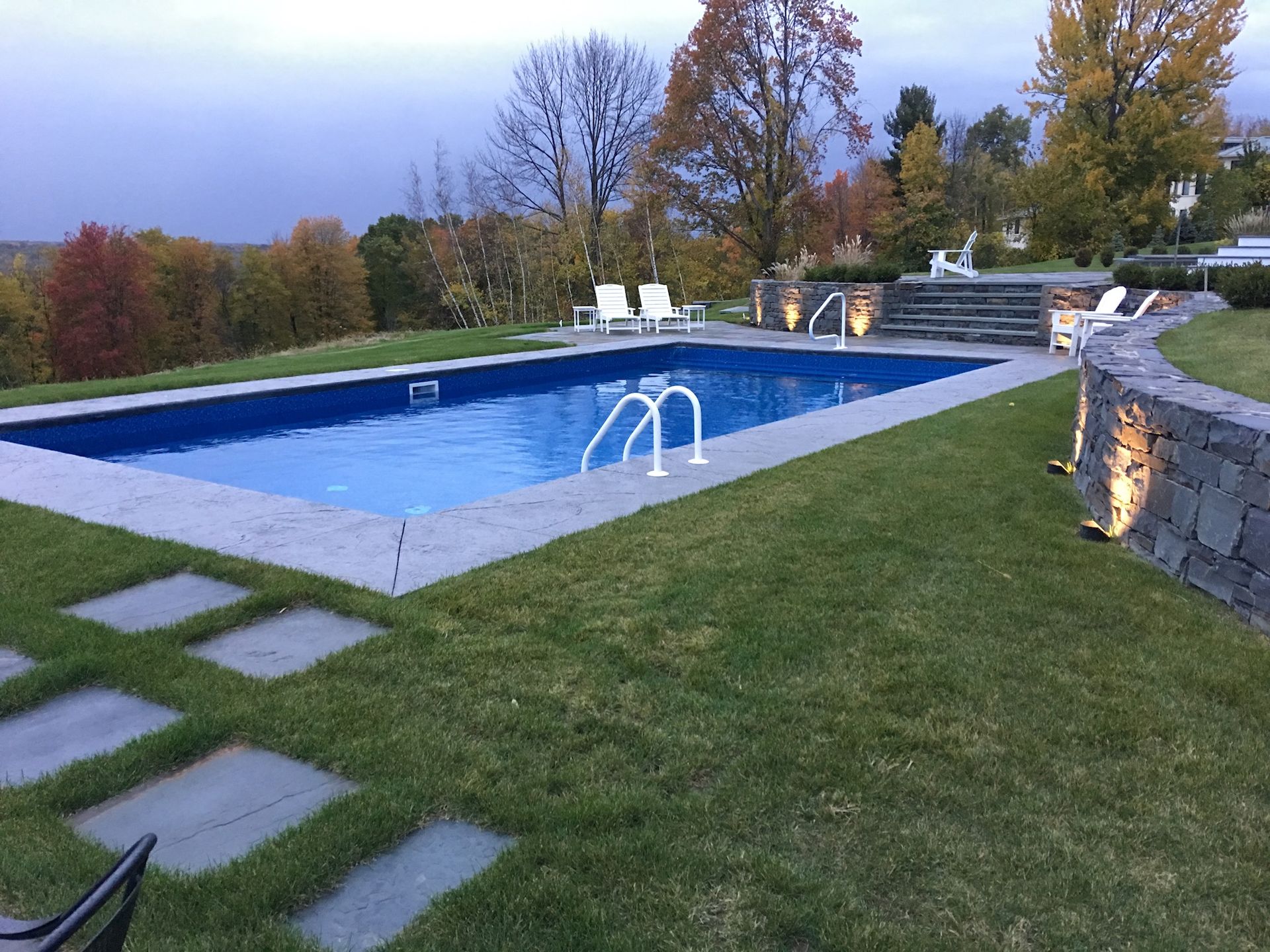 Rectangular pool surrounded by green grass and stone patio. Autumn trees in the background.