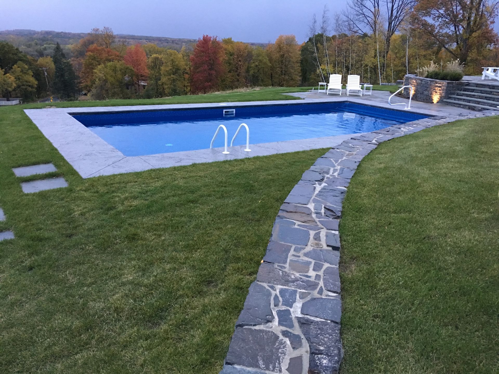 Pool in a grassy yard with stone pathway, steps, and autumn trees in the background.