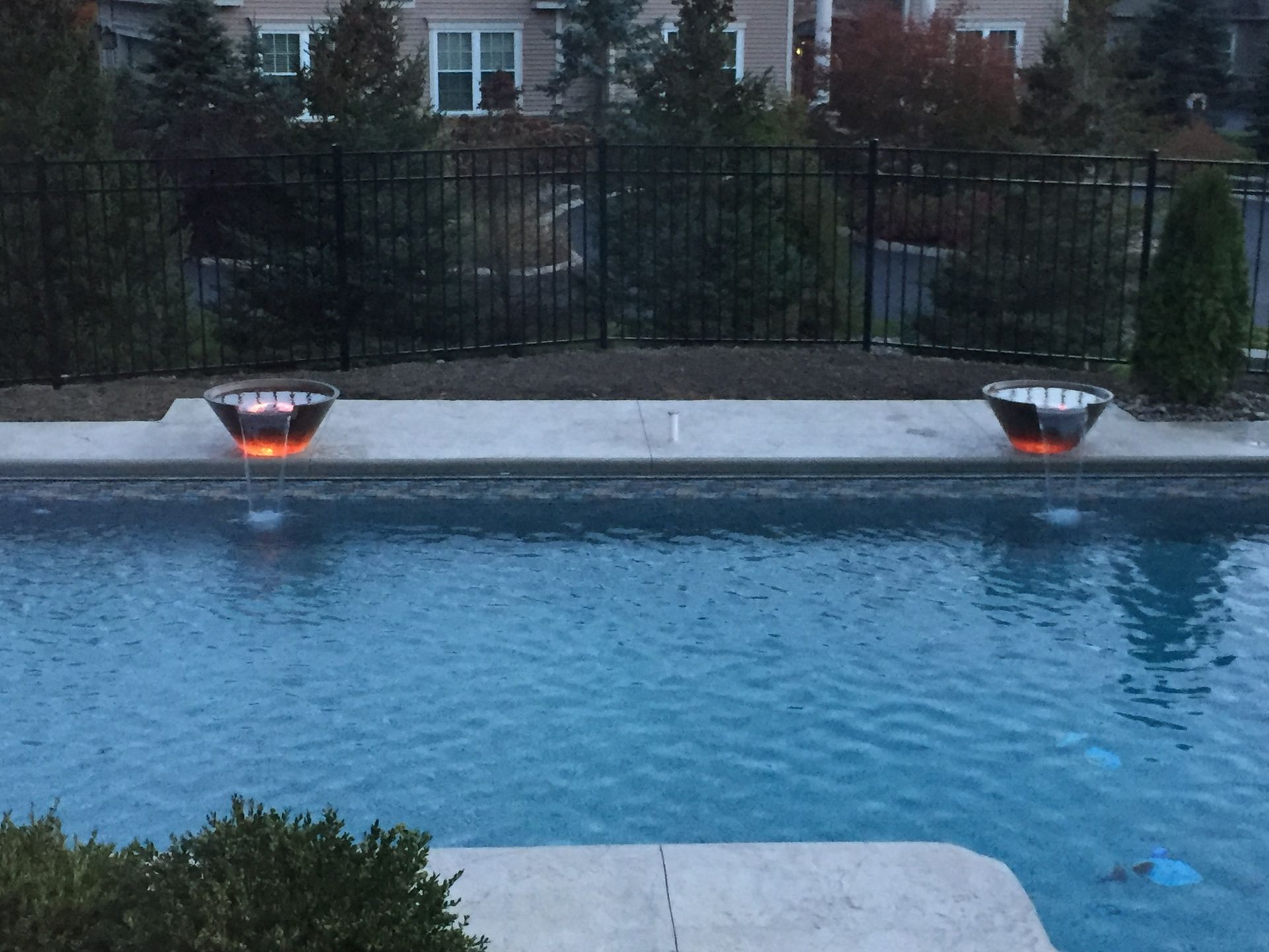 Pool with glowing fire bowls on the side. Evening, dark. Blue water, black fence, trees in background.