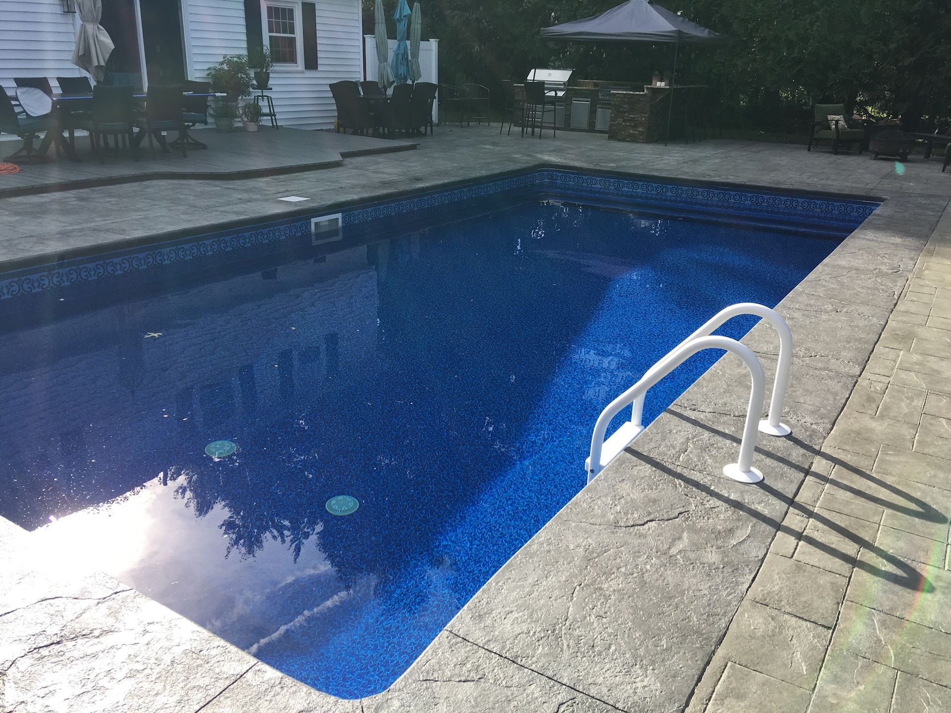 Rectangular blue-tiled pool with white ladder in a paved backyard; house and outdoor furniture visible.