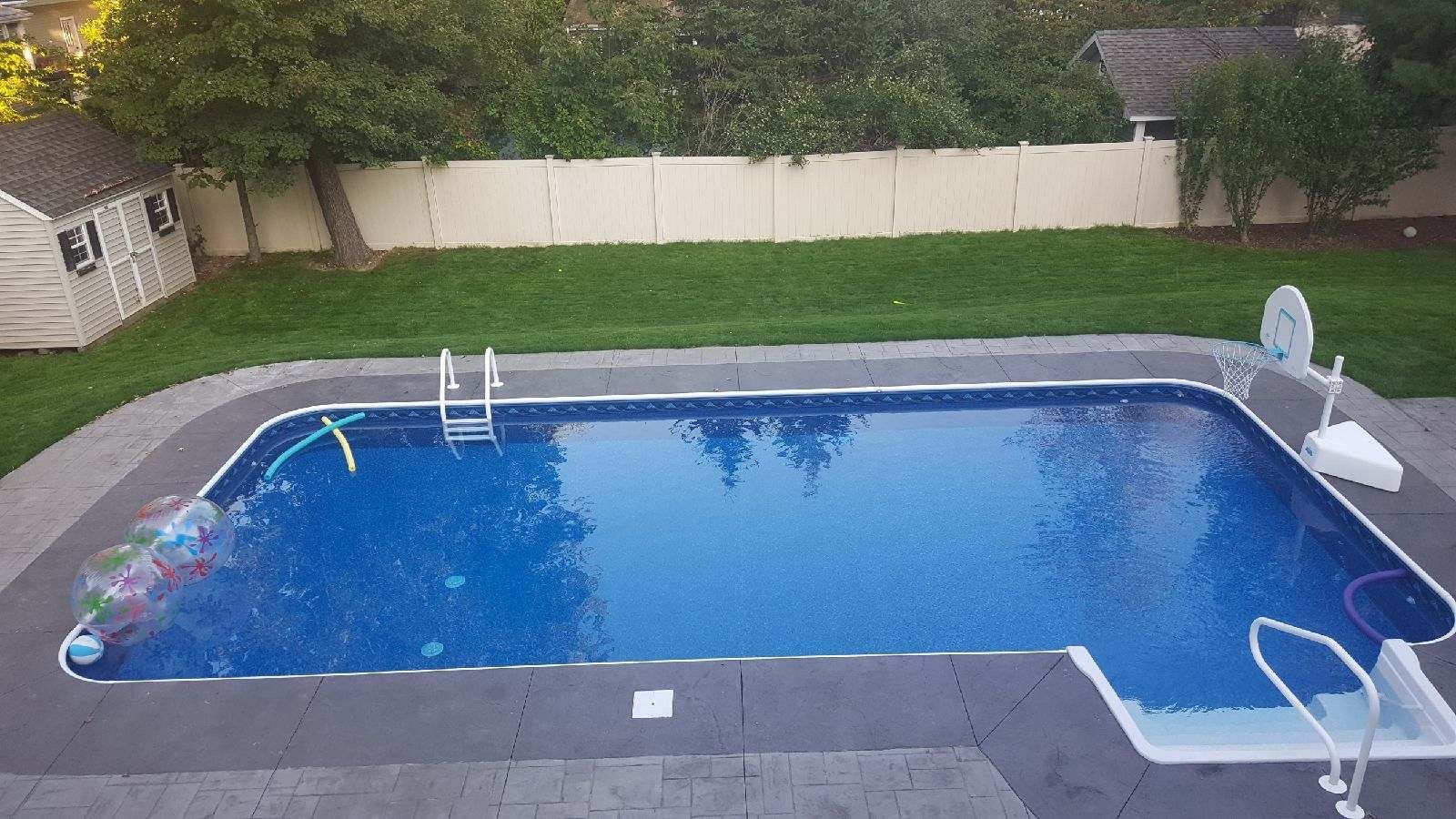 Rectangular in-ground pool surrounded by gray concrete, with green grass and white fence in the background.