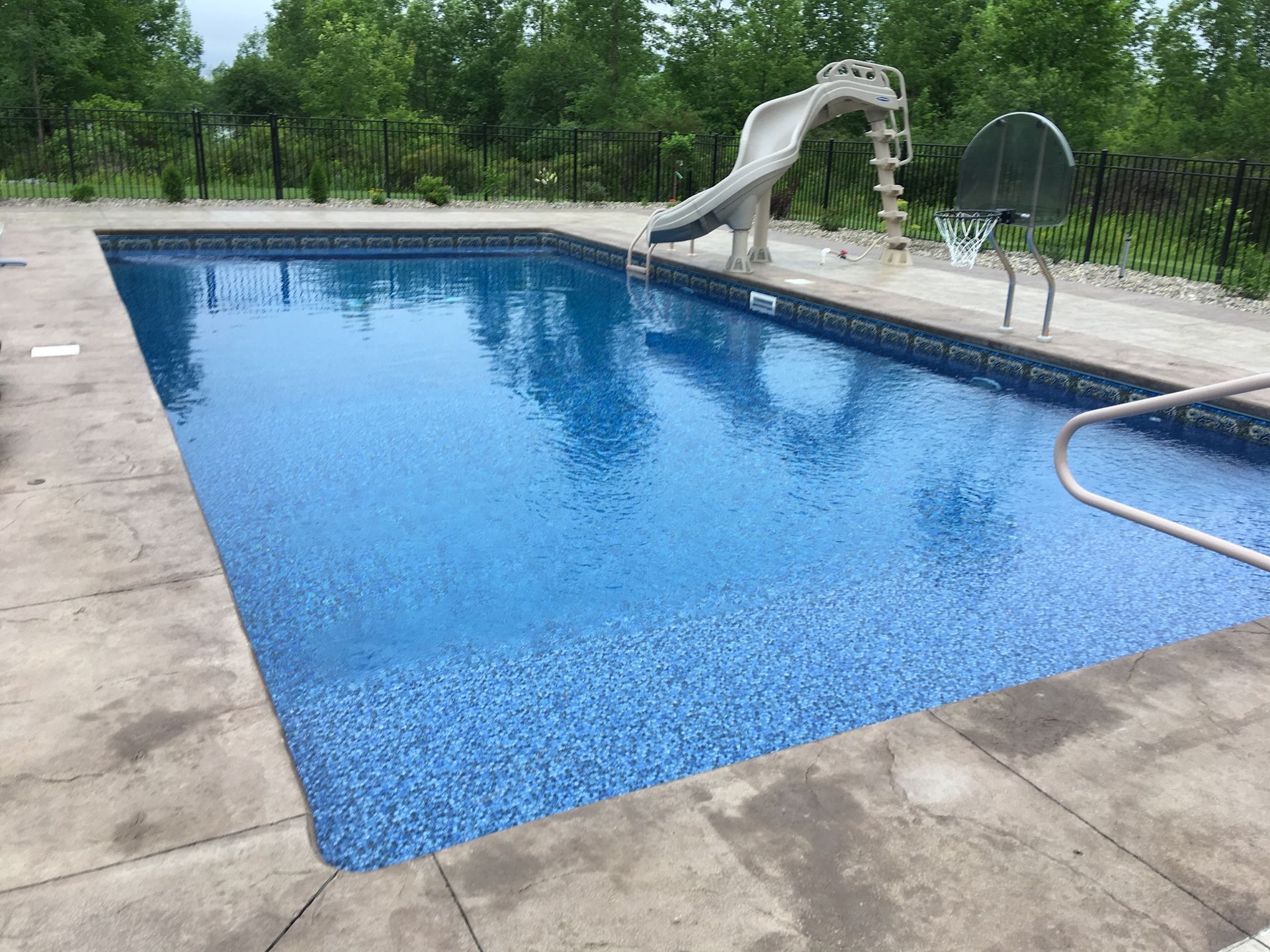 Rectangular in-ground pool with blue water and surrounding concrete. Slide and basketball hoop visible.