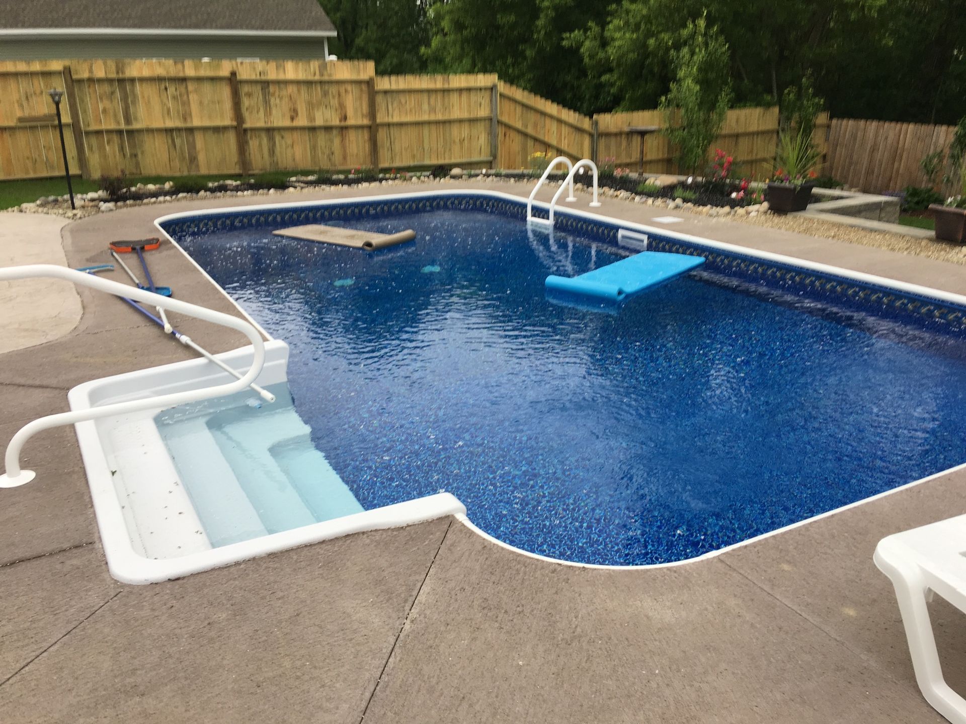 Rectangular in-ground swimming pool with blue water and surrounding concrete patio. A fence encloses the backyard.