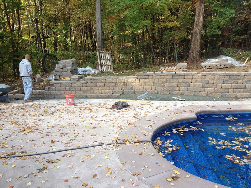 A person building a retaining wall near a pool with fall leaves, surrounded by trees.