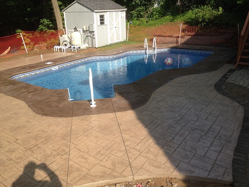 Swimming pool with stamped concrete patio and small white shed in background.