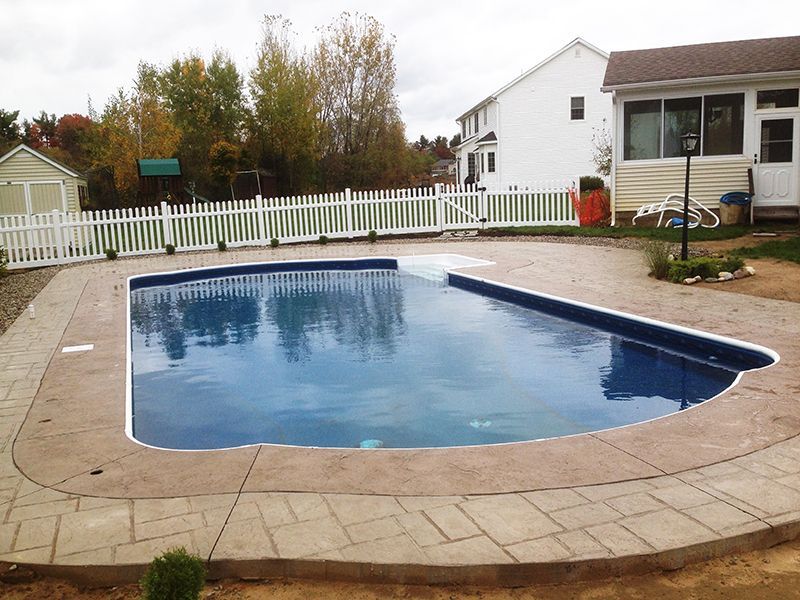 A backyard pool with blue water, surrounded by a brick patio and a white picket fence.