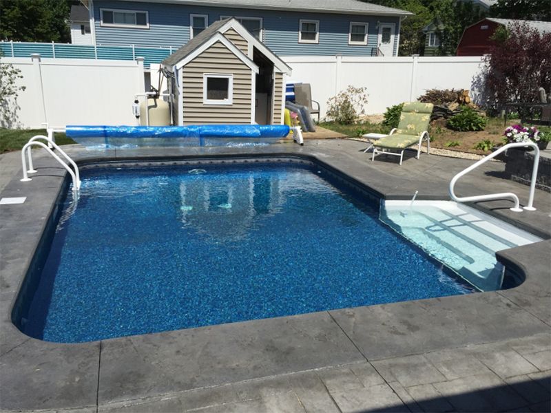 Swimming pool with blue water, concrete surround, and shed in the background. White fence and steps into pool.