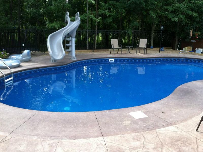 Swimming pool with slide, chairs, and surrounding concrete patio. Blue water.