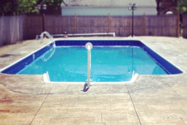 Blue pool in a concrete yard with a handrail. Wooden fence and house in the background.