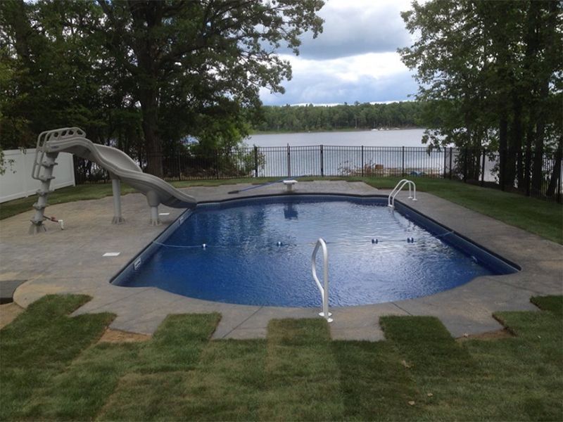 A rectangular swimming pool with a slide, fountain, and lake view behind a black fence.