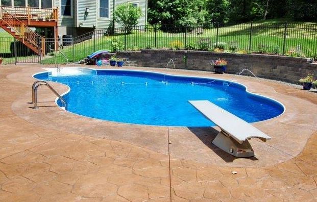 Swimming pool with a diving board, set in a brown-stamped concrete patio, adjacent to a house.