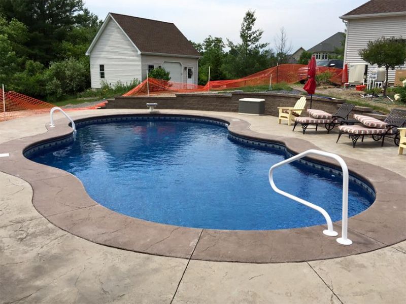 Pool with blue water and tan concrete surround; white safety rails; background includes a garage.
