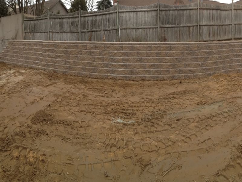 A modular block retaining wall backs a muddy area under a wooden fence.