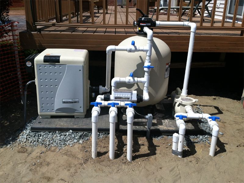 Pool filtration system with heater and sand filter, mounted on a gravel base near a wooden deck.