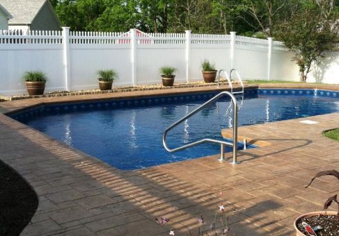 A rectangular pool with blue water, surrounded by brown brick patio, white fence, and potted plants.