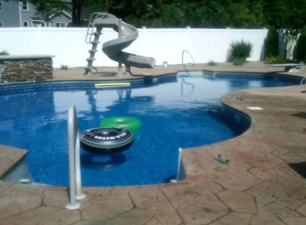 Pool with a slide, floats, and brown concrete deck in a backyard. Blue water, white fence.