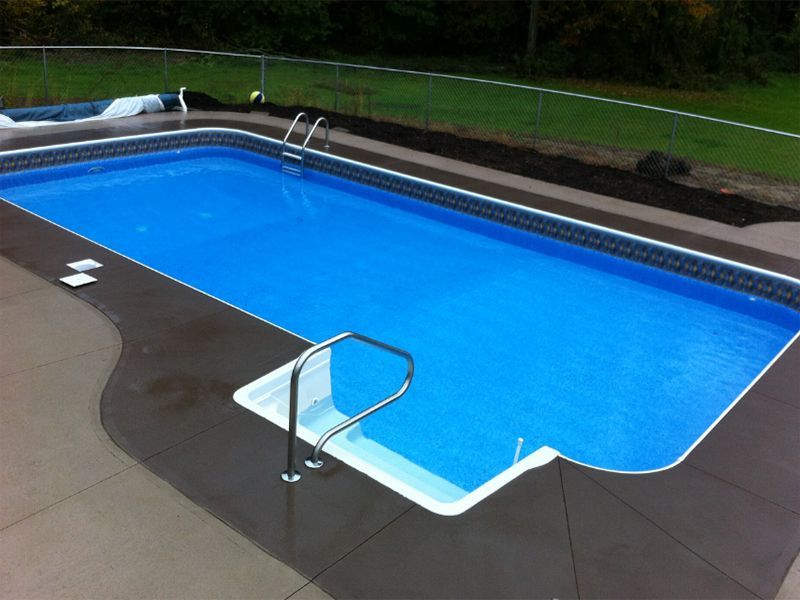 Rectangular blue swimming pool with dark gray concrete surround; in a backyard.