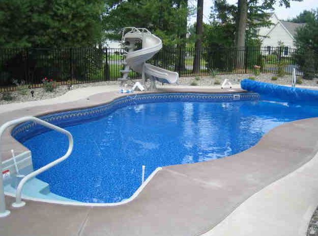 A curved in-ground swimming pool with a slide, surrounded by a concrete patio.