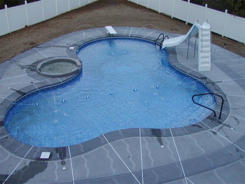 Overhead view of a kidney-shaped pool with a hot tub, slide, and diving board, surrounded by gray concrete.