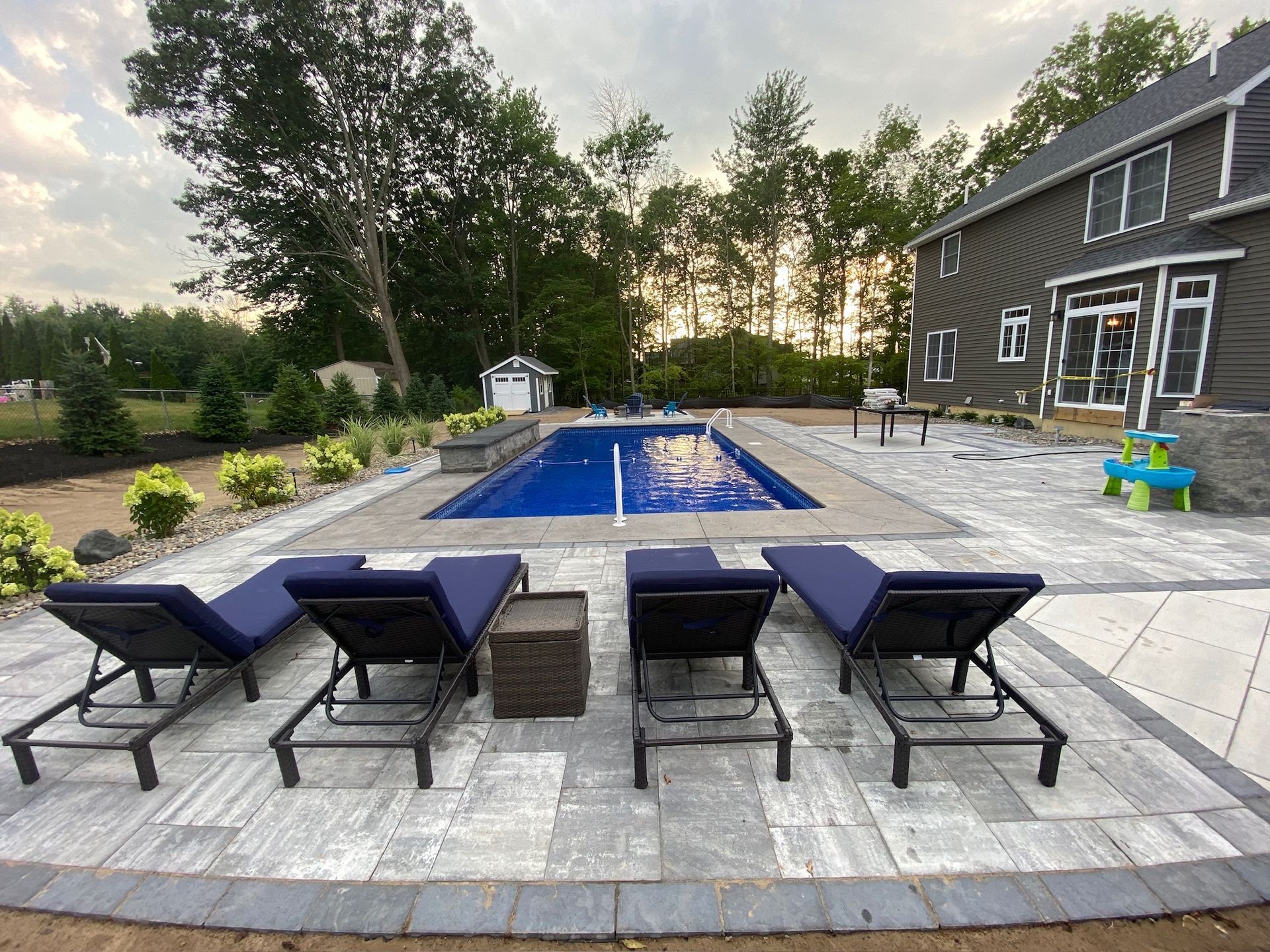 Four blue chaise lounges face a rectangular pool in a backyard patio setting with a house in the background.
