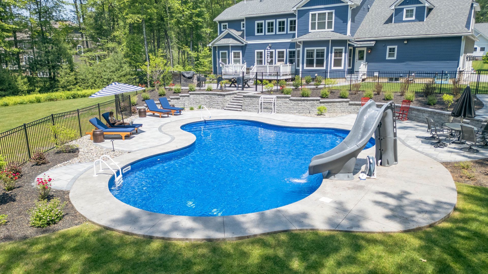 Blue pool with a slide in a backyard with a house in the background. Two lounge chairs by pool.