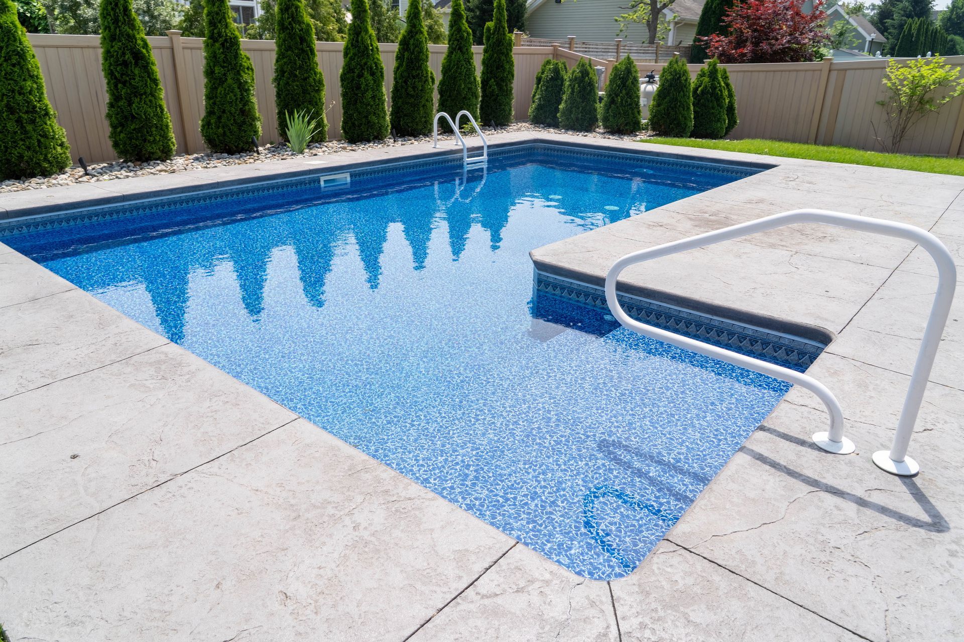 Rectangular swimming pool with blue water and white handrails, surrounded by concrete and landscaping.