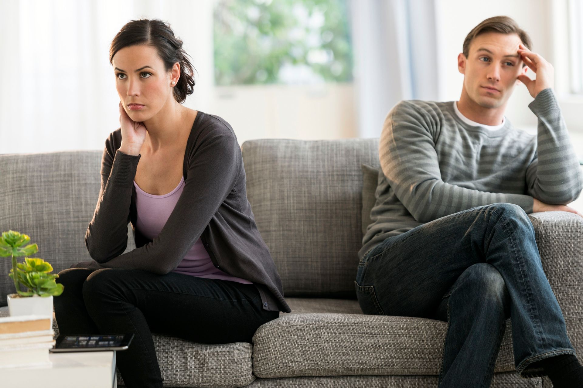 A couple sits apart on a grey sofa, looking away from each other with somber, pensive expressions.
