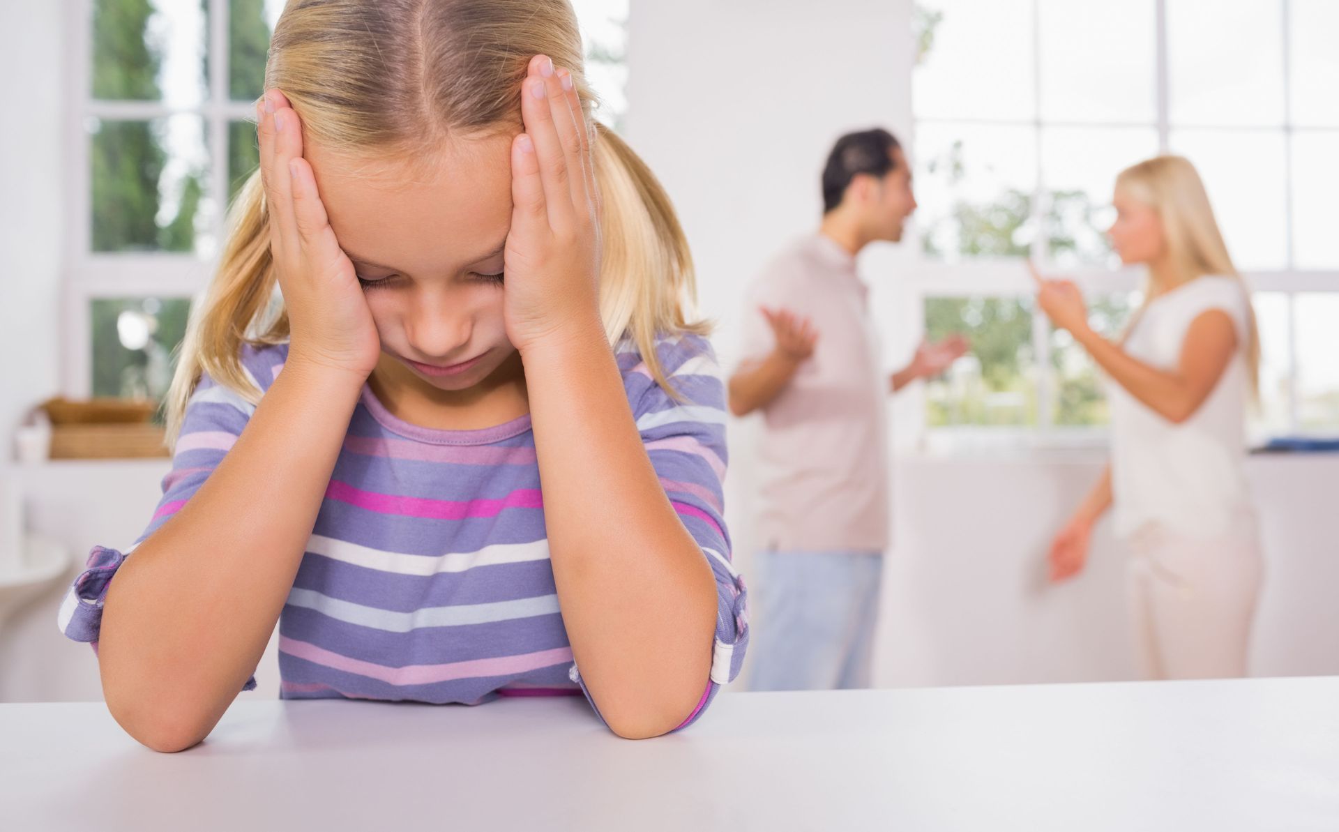A young child holds their head in distress while two adults argue in the background, out of focus.
