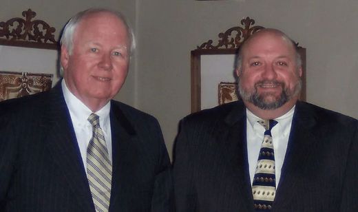 Two men in suits standing indoors, one in a striped tie and one in a patterned tie.