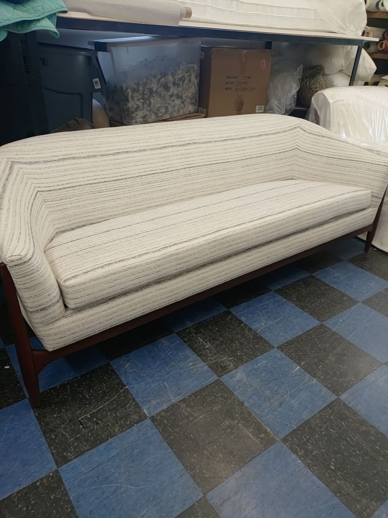 A mid-century modern sofa with cream-colored patterned fabric and dark wooden legs in a workshop with a tiled floor.